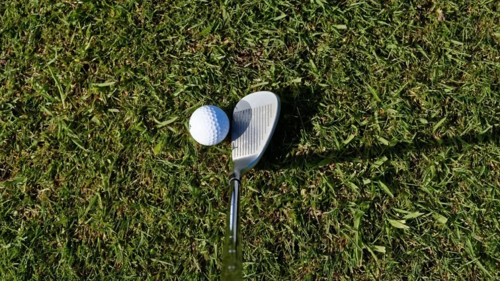A golf wedge lining up a shot of a white golf ball on green grass and sand