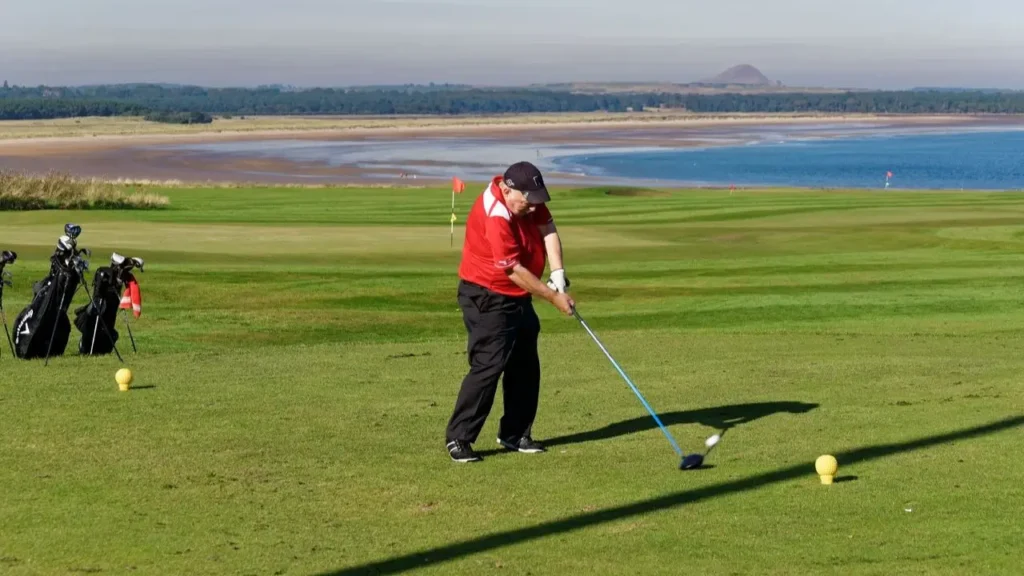 A golfer wearing a red golf shirt hitting a golf ball with a golf club driver on a golf course green