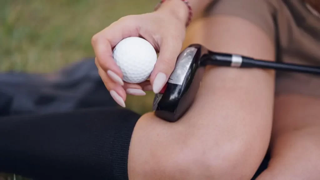 A female golfer sitting with a golf club on her leg whilst holding a white golf ball