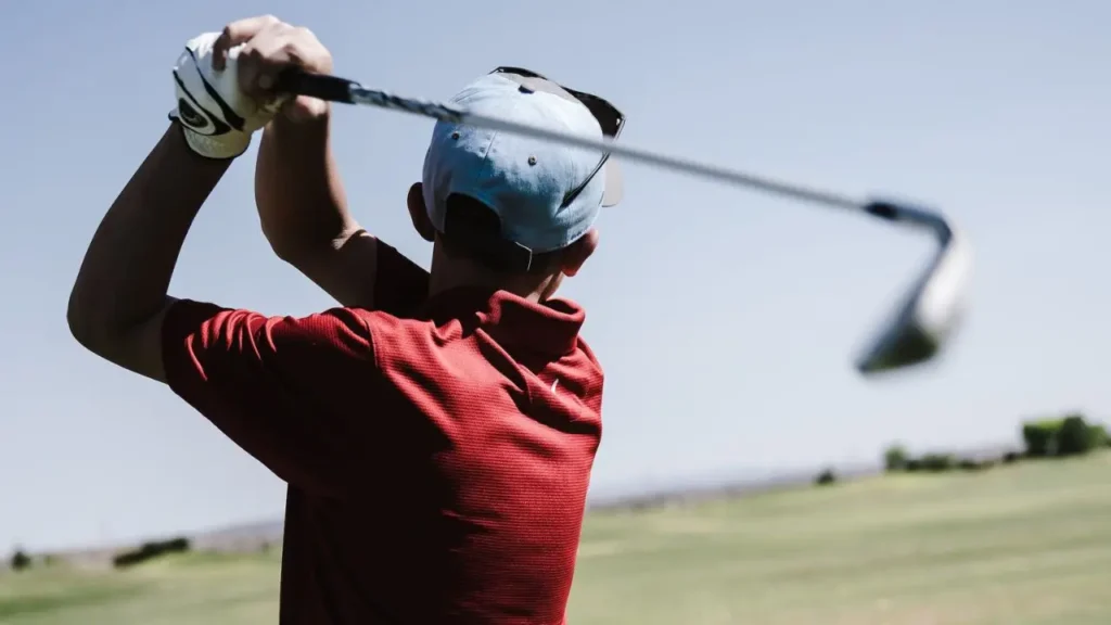 A golfer wearing a red golf shirt swinging a golf iron wearing a grey cap
