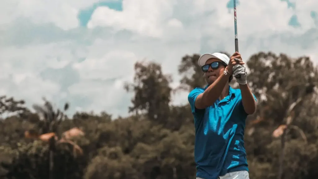 A golfer wearing a blue shirt and white cap striking a white golf ball on a golf course