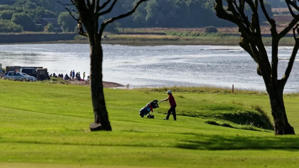 A golfer in the distance of a golf course with a river behind pulling a golf trolley bag